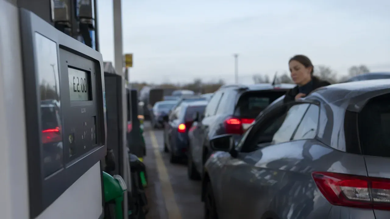 Close-up of a UK fuel pump display showing £2.00/litre with a queue in background